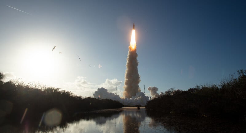 A rocket launches into a clear blue sky, leaving behind a trail of smoke and flames. Birds fly nearby, and the scene is reflected in the calm water below, with vegetation framing the view.