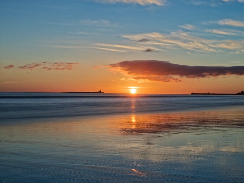 A serene beach at sunset with the sun low on the horizon, casting a warm orange glow over the water and wet sand. Clouds are scattered in the sky and the scene is calm and peaceful.