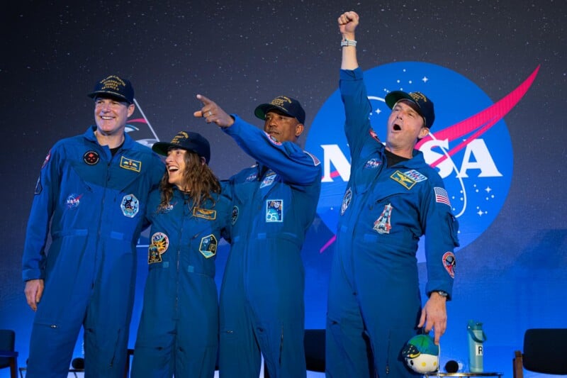 Four astronauts in blue flight suits stand in front of a NASA logo, smiling and celebrating; one raises a fist triumphantly while others pose beside him on a stage.