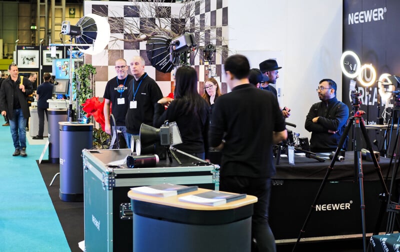 A group of people stands at a photography equipment booth with lights, cameras, and displays on tables. The booth displays the Neewer brand, and attendees are engaged in conversation.