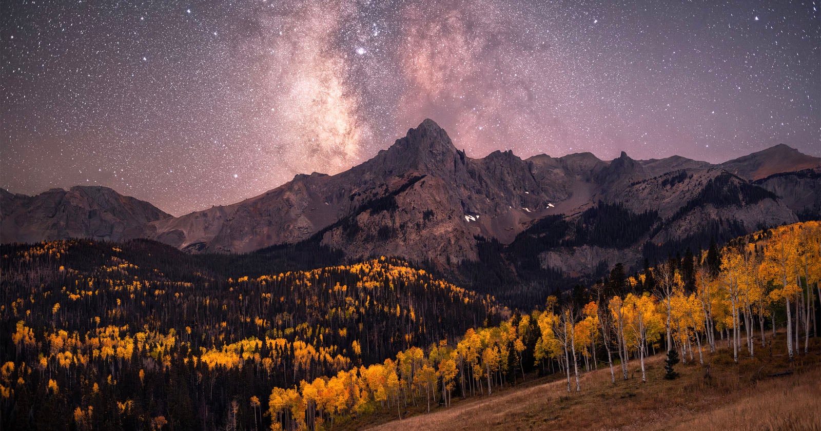Snow-capped mountain peak under a star-filled night sky with the Milky Way visible; yellow autumn trees cover the hills in the foreground.