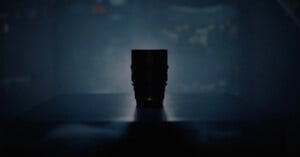 A camera lens is silhouetted against a dark, blurred background, sitting on a flat surface with soft, moody lighting highlighting its outline.