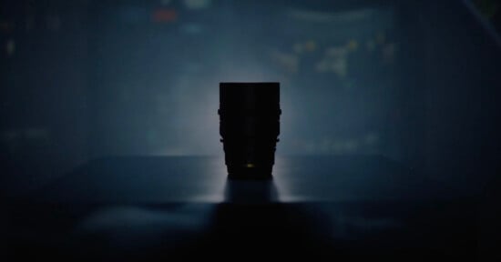 A camera lens is silhouetted against a dark, blurred background, sitting on a flat surface with soft, moody lighting highlighting its outline.