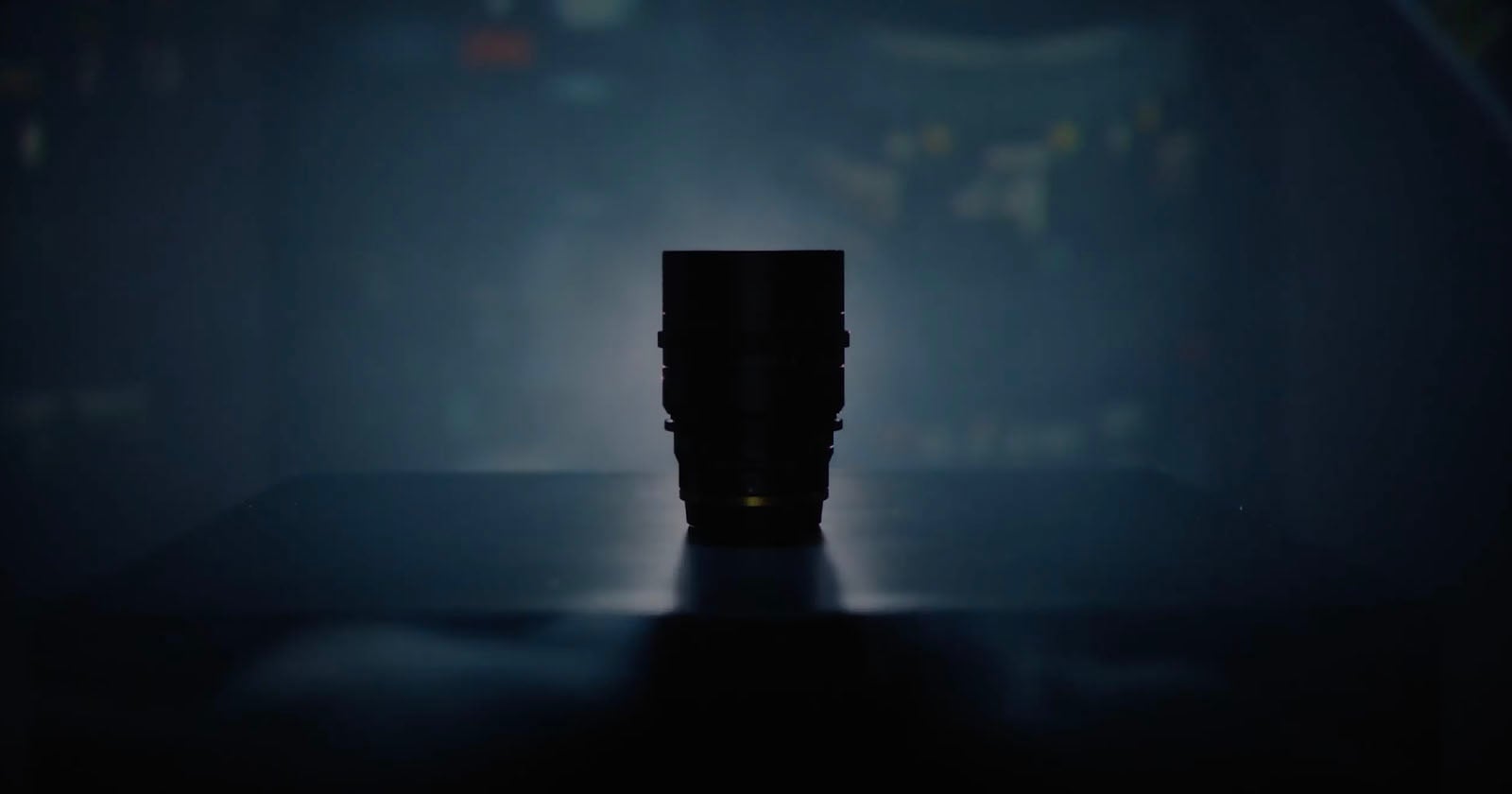 A camera lens is silhouetted against a dark, blurred background, sitting on a flat surface with soft, moody lighting highlighting its outline.