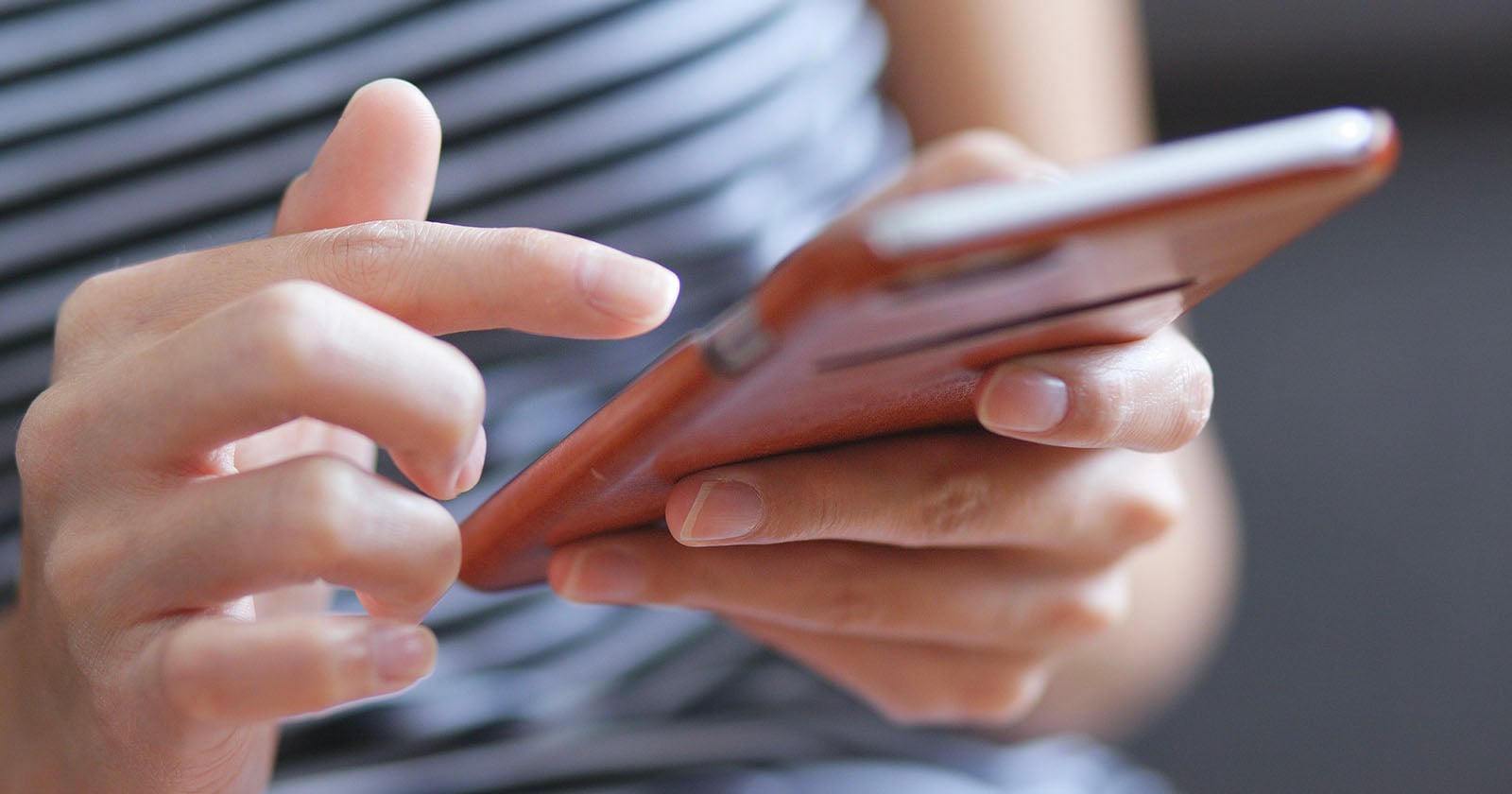 Close-up of a person using a smartphone, holding it in one hand and tapping the screen with the other. The person is wearing a striped shirt.