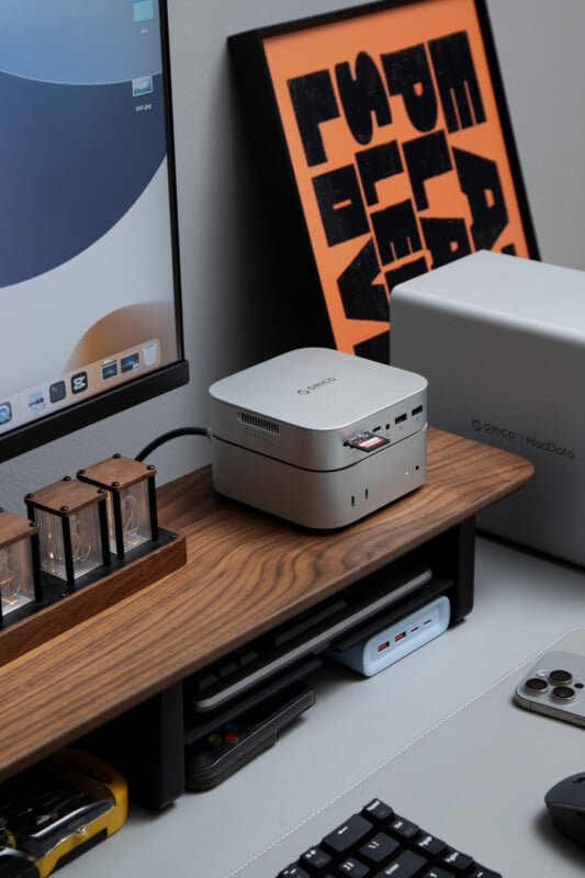 A minimalist desktop setup with a monitor, a small silver mini PC, decorative clock tubes, a keyboard, and an orange and black framed poster leaning against the wall.