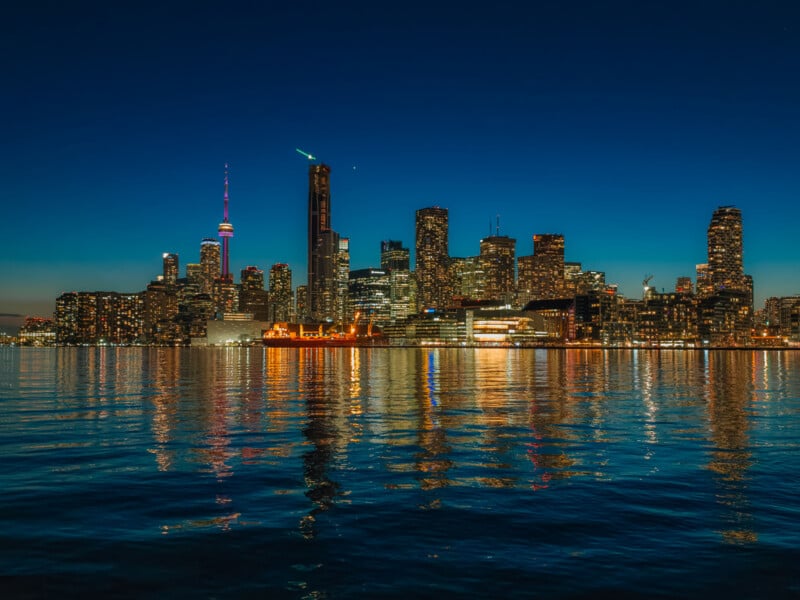 A brightly lit city skyline at dusk is reflected on calm water, with a prominent tower featuring colored lights standing among tall buildings against a deep blue sky.