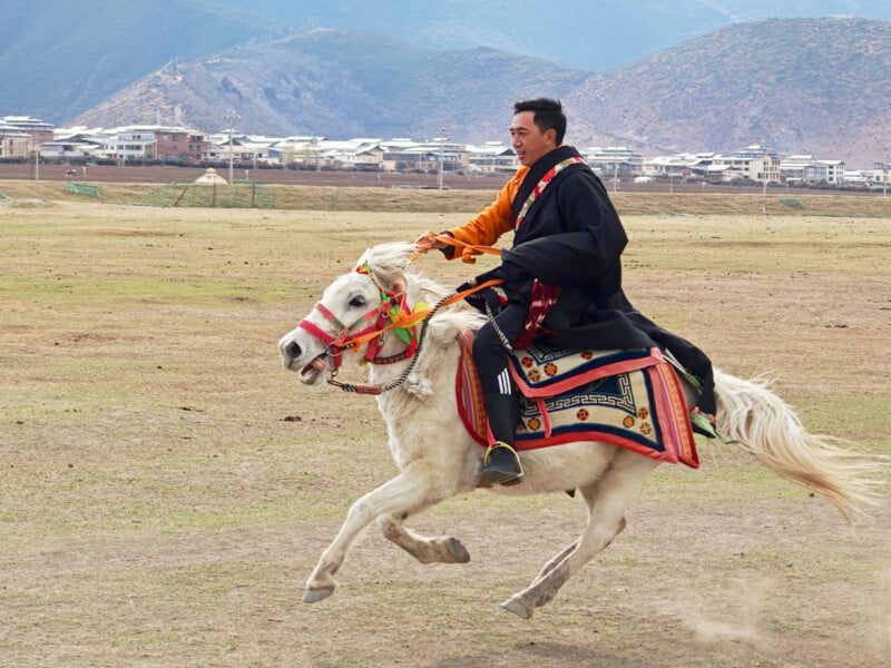 A man wearing traditional clothing rides a white horse across an open grassy field, with mountains and a village visible in the background.