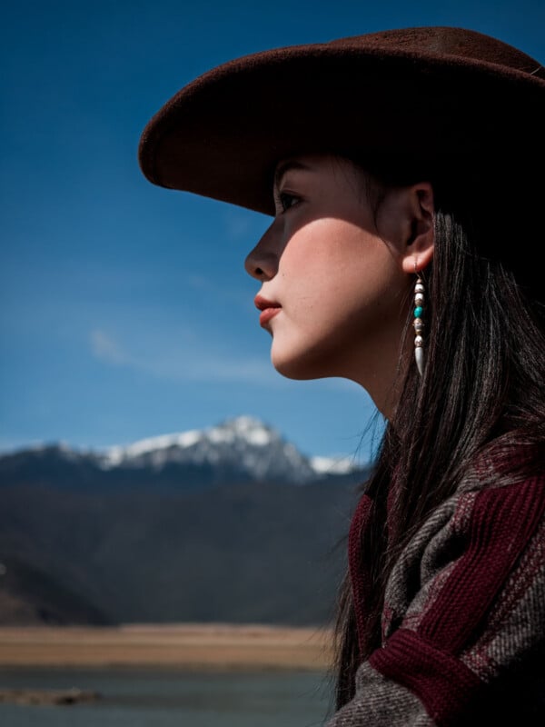 A woman in profile wearing a wide-brimmed hat and beaded earring is outdoors with snow-capped mountains and a blue sky in the background.