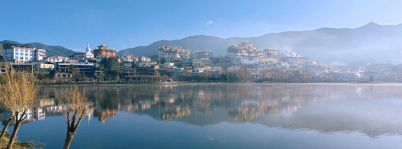 A panoramic view of a town with traditional-style buildings and temples on a hillside, reflected in a calm lake, with mountains and blue sky in the background.