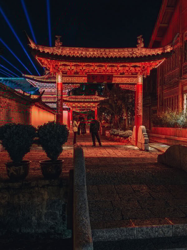 A traditional Chinese gate illuminated with red lights at night; people stand underneath the archway on a stone path, with blue light beams shining in the sky and ornate buildings in the background.