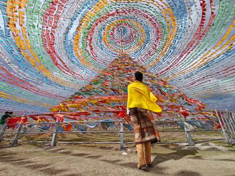 A person in a yellow shawl stands before a large cone-shaped structure adorned with colorful, swirling prayer flags, set outdoors under a sky filled with more vibrant flags.