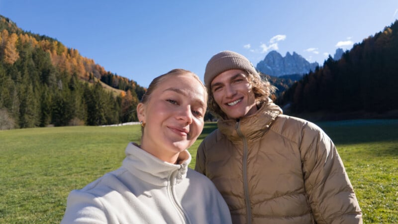Two people in warm jackets take a selfie outside on a sunny day, standing in a grassy field with forested hills and snow-capped mountains in the background.