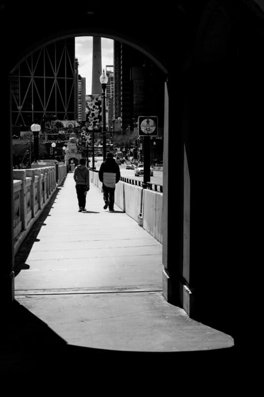 Two people walk along a city sidewalk, framed by the shadowy entrance of a tunnel. Tall buildings, streetlights, and a busy street are visible in the background on a sunny day. The image is in black and white.