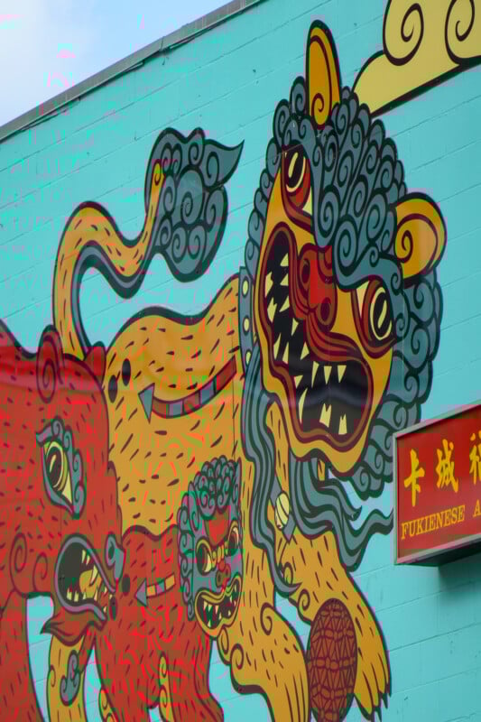 A colorful mural featuring stylized, fierce-looking lion-like creatures with swirling manes and exaggerated facial features. A red and yellow sign with Chinese characters and "FUKENESE" is visible on the right.