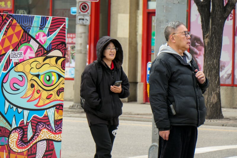 Two people in winter clothing stand on a city sidewalk near a colorful, abstract street art box. One holds a phone, and the other wears glasses and faces the street. Buildings and street signs are visible in the background.