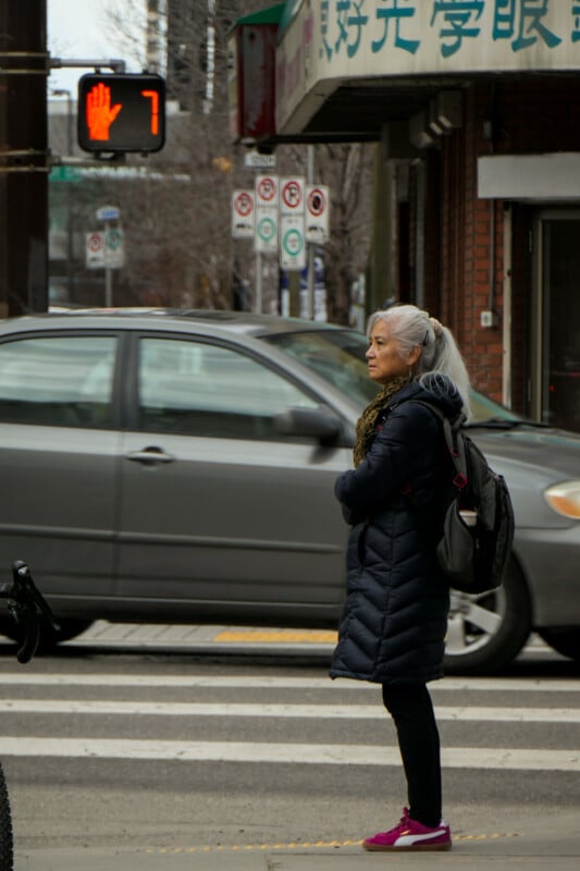 An older woman with gray hair, a dark coat, backpack, and pink shoes stands at a crosswalk as cars pass by. A red hand pedestrian signal and a countdown timer display the number 7 above the street.