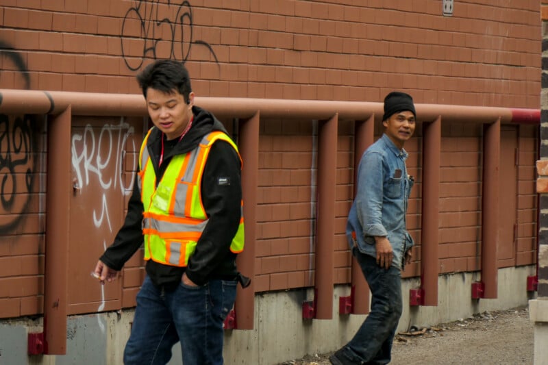 Two men walk along a sidewalk by a brick wall with graffiti. One wears a reflective safety vest and black hoodie, the other a denim jacket and black hat. Both appear relaxed and are looking in different directions.