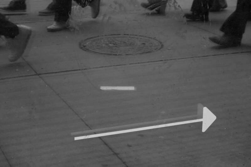 A group of people walking on a sidewalk, seen from the knees down. A white arrow and dash are marked on the pavement, pointing to the right. The image is in black and white.