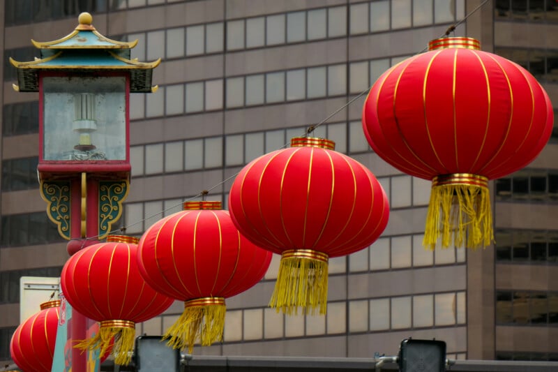 Red Chinese lanterns with gold accents hang in a row on a wire next to an ornate street lamp, set against the backdrop of a modern city building with many windows.