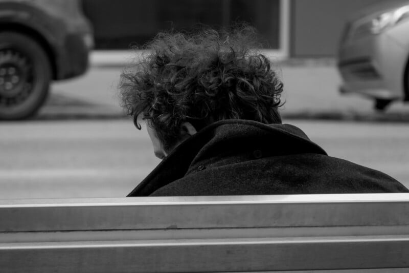 A person with curly hair, seen from behind, wears a dark coat while sitting on a bench by the street. The image is in black and white, with parked cars blurred in the background.