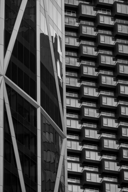 Black and white photo of two modern high-rise buildings; one has a geometric glass facade with diagonal lines, and the other features a repetitive pattern of small, protruding balconies.