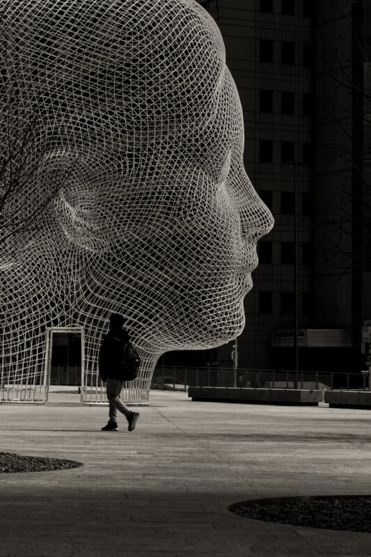 A person walks past a large wire mesh sculpture of a human head in an urban plaza, with tall buildings in the background. The image is in black and white, highlighting the sculpture’s intricate structure.