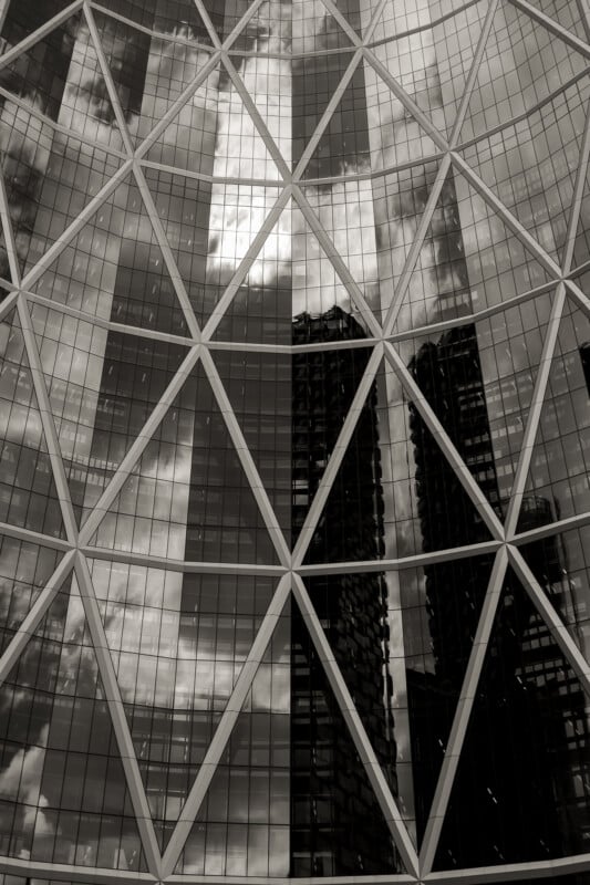 A close-up view of a glass building facade with a geometric metal grid pattern, reflecting nearby skyscrapers and clouds in the windows. The image is in black and white.