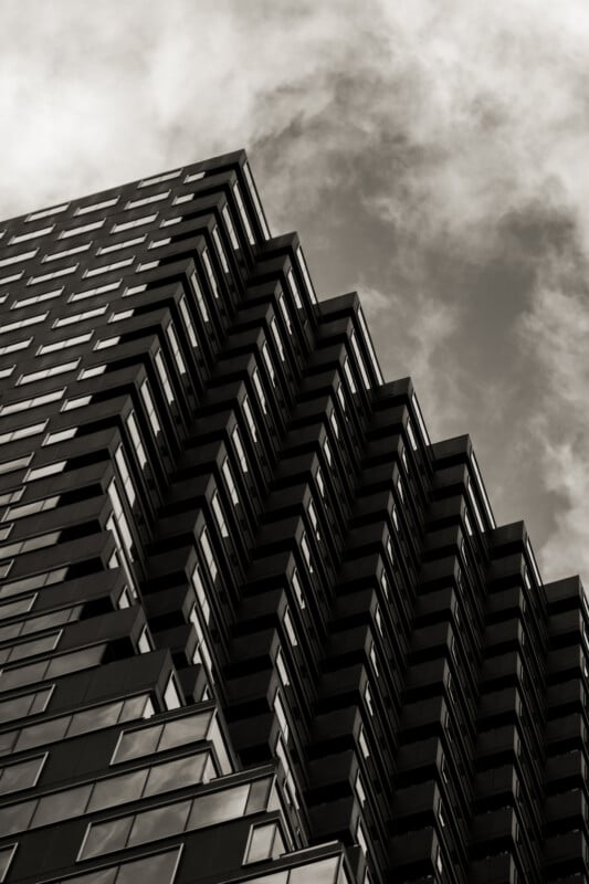 A dramatic, upward view of a modern high-rise building with staggered, geometric balconies against a cloudy sky, rendered in black and white.