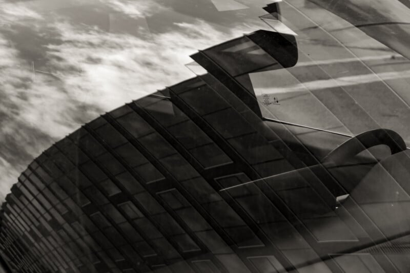 Black and white photo showing reflections of clouds and a building with many windows on a glass surface, creating an abstract, layered effect. The angles and lines blend together, making it hard to distinguish the objects.