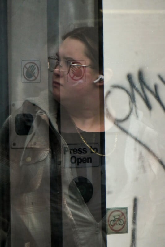 A person with glasses and earbuds looks to the side behind a glass panel covered with reflections, stickers, and graffiti. They wear a plaid jacket, a gold chain, and a shirt reading "Press to Open.