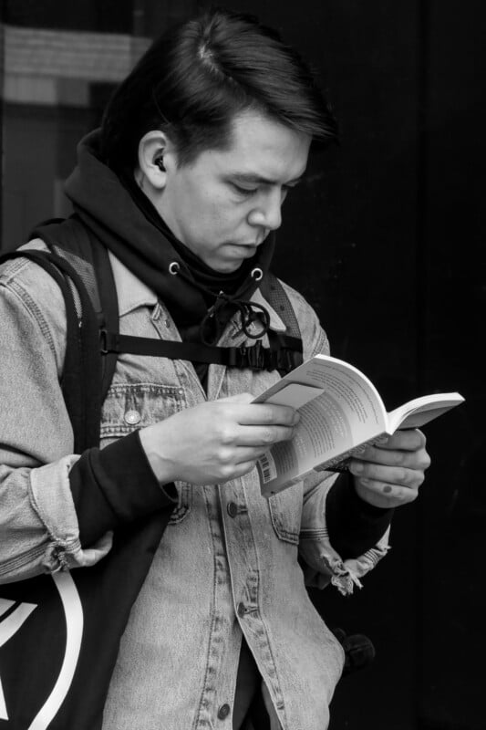 A person with straight dark hair, wearing a denim jacket over a hoodie, a backpack, and earbuds, is standing outdoors and reading a book. The image is in black and white.