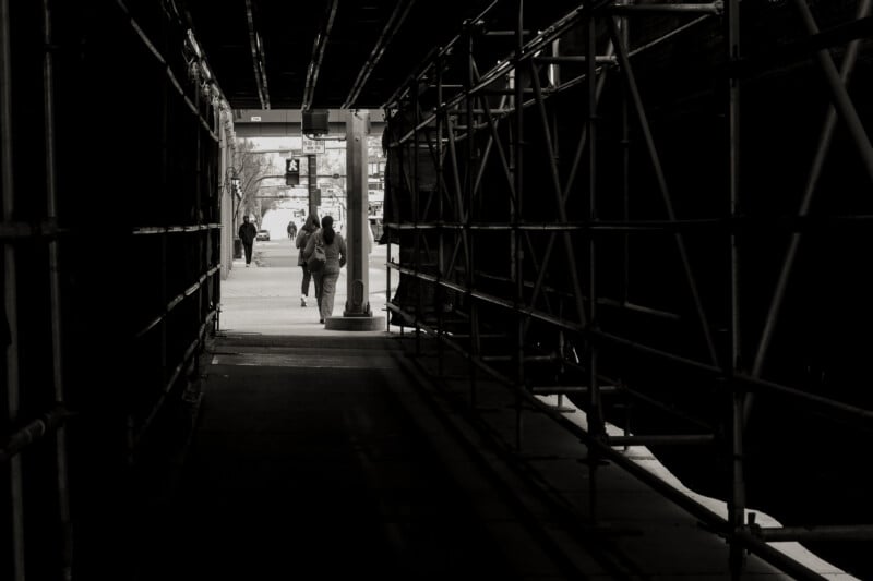 Two people walk away from the camera through a dark, scaffolded tunnel toward a brighter street scene outside, with other figures and a pedestrian crossing in the distance.