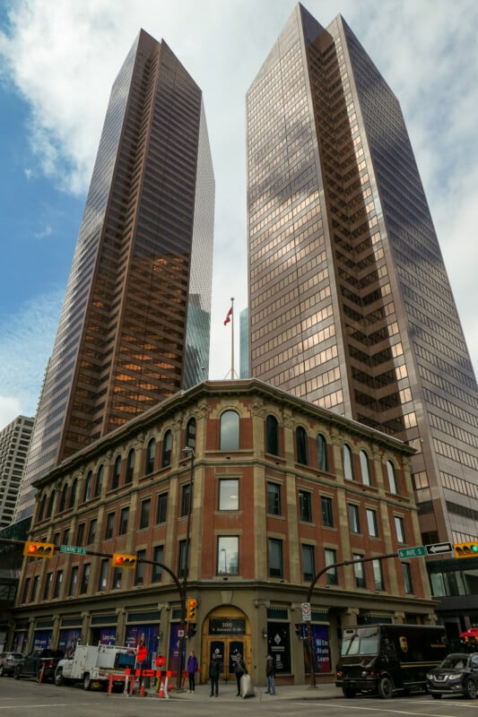 A historic three-story brick building stands at a street corner, dwarfed by two modern glass skyscrapers behind it. People walk on the sidewalk, and vehicles are stopped at the intersection under a cloudy sky.