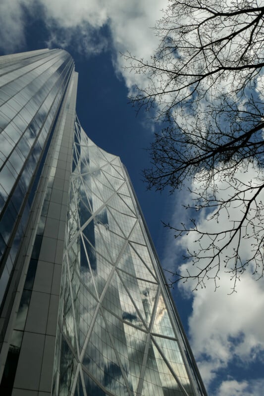 A modern glass skyscraper rises toward a partly cloudy sky, its reflective surface capturing clouds. Bare tree branches appear on the right side of the image.
