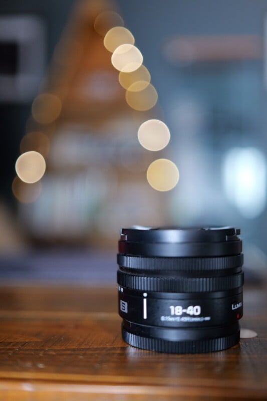 A Lumix 18-40mm camera lens sits on a wooden surface, with blurred, circular bokeh lights and an out-of-focus background creating a soft, artistic effect.