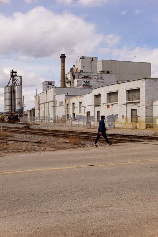 A person in dark clothing walks across train tracks in front of a large, weathered industrial building with silos and a tall smokestack under a partly cloudy sky.
