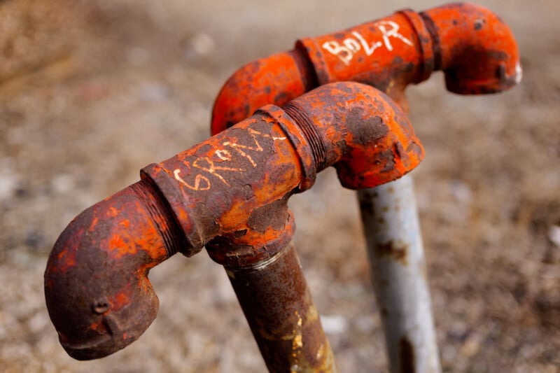 Two rusted, red-painted metal pipes with elbow joints stand side by side outdoors. Both pipes have white graffiti on top and show signs of weathering and corrosion. The background is blurred.