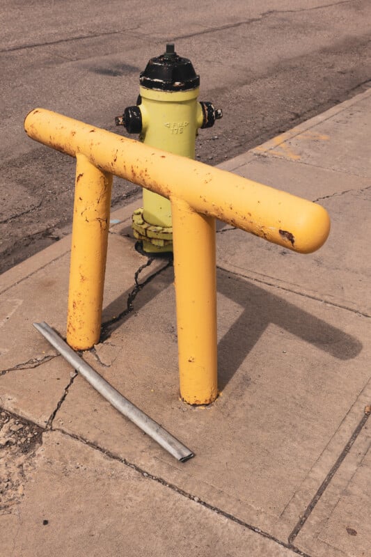 A yellow metal guardrail surrounds a green fire hydrant on a concrete sidewalk next to a street. A detached metal pipe lies on the ground nearby. The scene shows signs of wear and rust.
