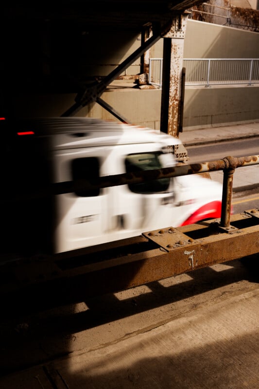 A white and red vehicle moves quickly under a rusty overpass, creating a motion blur effect. Sunlight and shadows highlight the industrial structure and road above.
