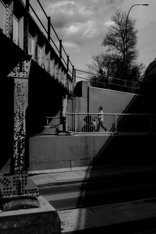 A person walks on a pedestrian bridge under a railway track in a city. Shadows and sunlight create strong contrasts in this black and white photo. A bare tree and cloudy sky are in the background.