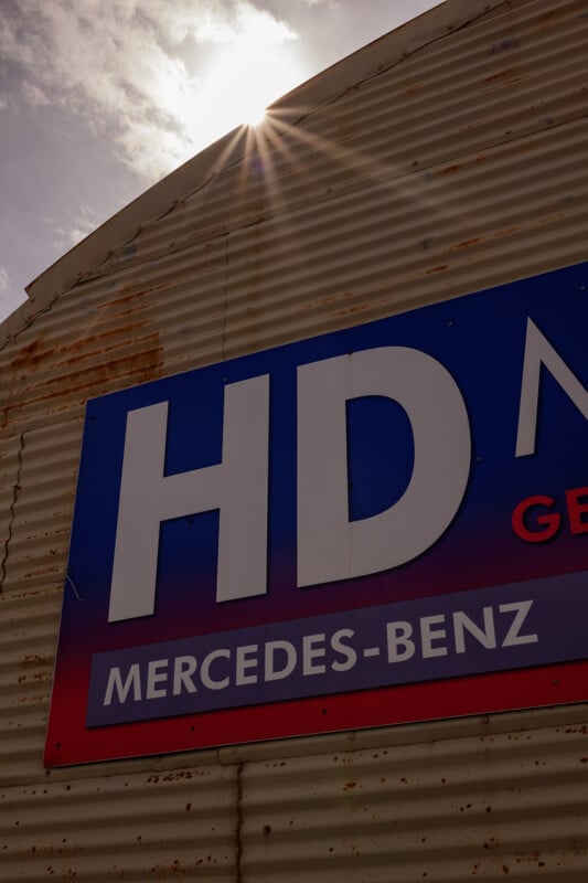 A corrugated metal building with a partial sign reading "HD" and "MERCEDES-BENZ" under sunlight, with the sun peeking over the roof edge and a partly cloudy sky.