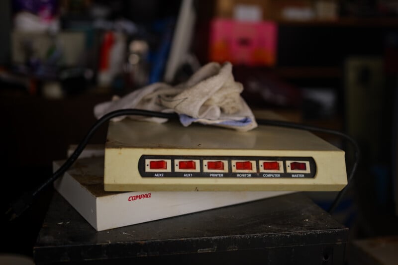 A vintage switch box with six red switches labeled AUX2, AUX1, PRINTER, MONITOR, COMPUTER, and MASTER sits on top of a white Compaq device, with a cloth draped over it in a cluttered room.