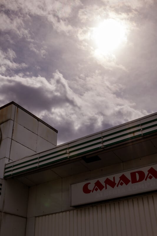 A cloudy sky with the sun shining through is seen above the corner of a white building. Part of a sign reading "CANADA" is visible on the building's facade.