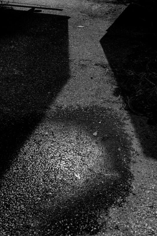 A black-and-white photo shows a rough, wet patch on a paved surface, illuminated by light. Tall, angular shadows from nearby objects create dramatic contrasts on the ground.