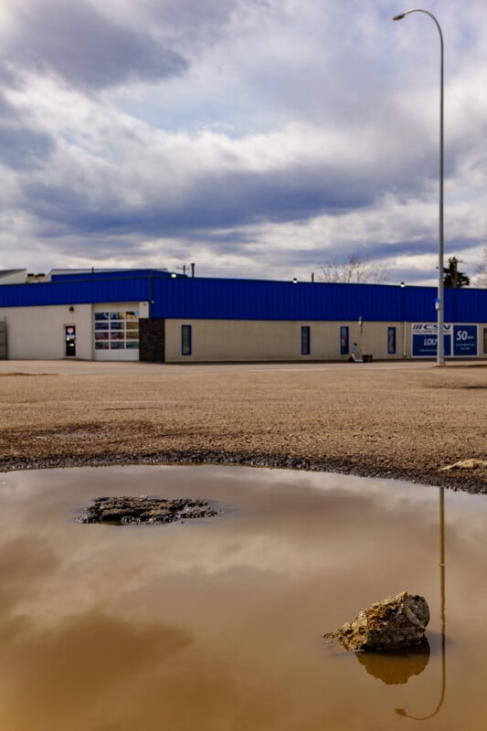 A muddy puddle with a rock in the foreground reflects the cloudy sky and a lamppost. In the background, there's a building with a blue roof and a mostly empty gravel lot.
