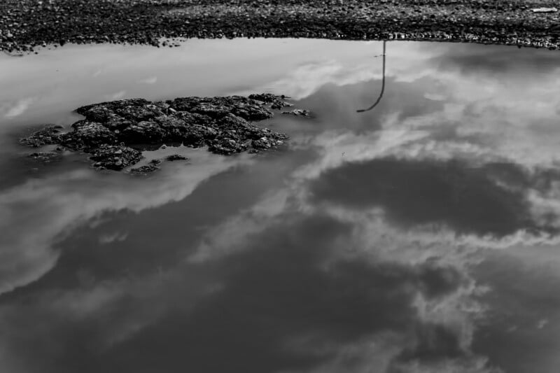 A black and white photo shows a puddle on asphalt reflecting clouds and a streetlamp, with a patch of rough, textured surface emerging from the water.