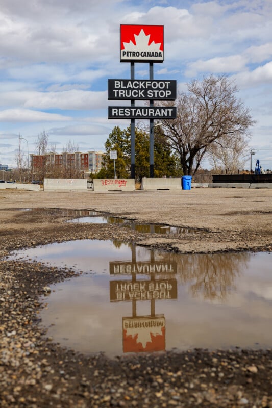 A Petro-Canada sign above a Blackfoot Truck Stop Restaurant sign is reflected in a large puddle on an empty, rough parking lot with cloudy skies and bare trees in the background.