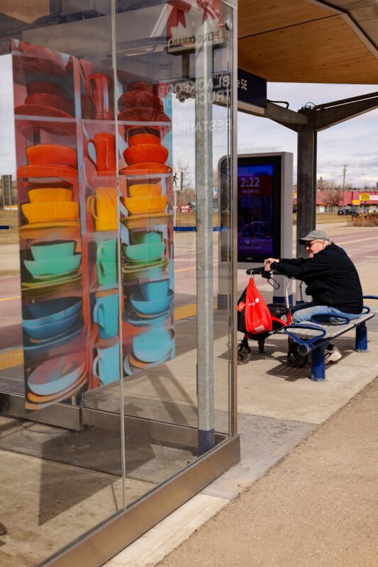 An older man sits on a bench at a bus stop, holding a red shopping bag. The bus shelter’s glass wall features a colorful image of stacked cups and bowls in various bright colors.