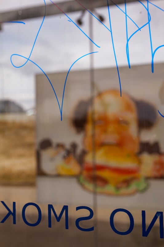 A window with blue graffiti and the reversed text "SMOK" partially visible. Behind the glass, there is a blurred sign showing a cartoon character holding a large burger. The background appears to be outdoors.